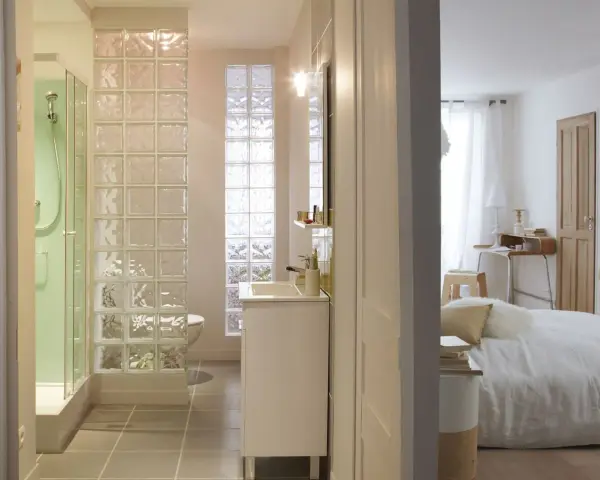 Salle de bain moderne avec douche, lavabo et toilettes, séparée de la chambre par un mur en brique de verre.