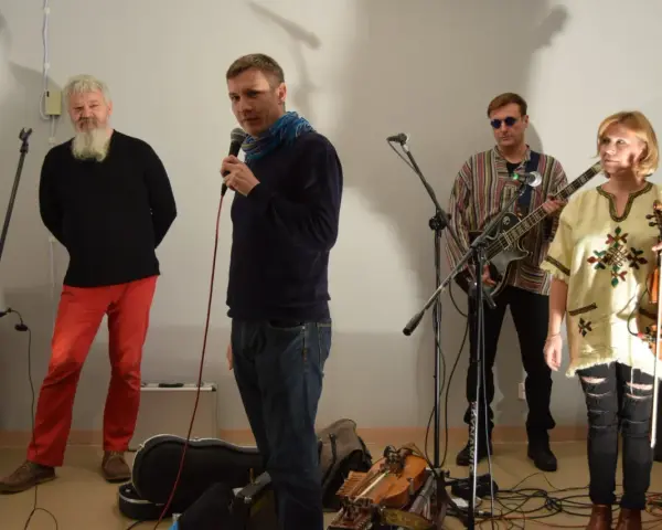 Orkiestra Świętego Mikołaja przygotowuje się do występu. Wokalista z mikrofonem, gitarzysta, skrzypaczka i brodaty mężczyzna w czerwonych spodniach.