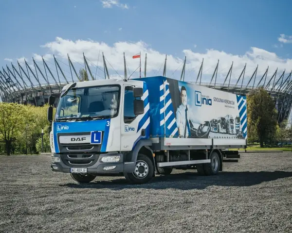 Ciężarówka szkoleniowa z literą "L" na masce, gotowa do nauki jazdy kat. C. W tle stadion.