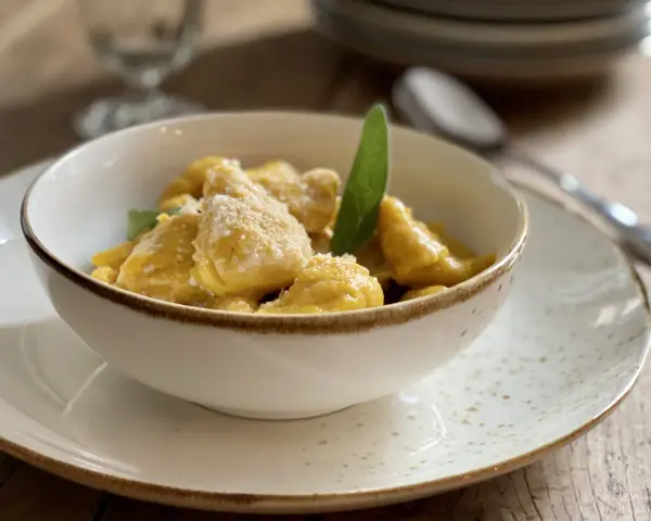 Gnocchi di zucca dorati e soffici, conditi con parmigiano e una foglia di salvia, serviti in una ciotola bianca su un piatto maculato.