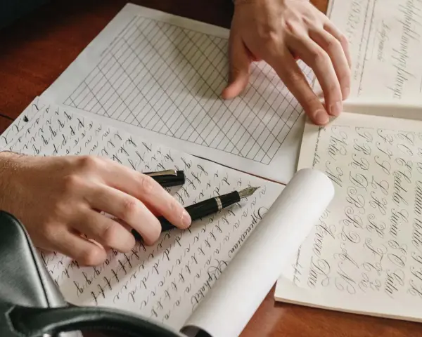 Manos practicando caligrafía con una pluma para colorear, practicando letras y formas en papel cuadriculado y con guías.