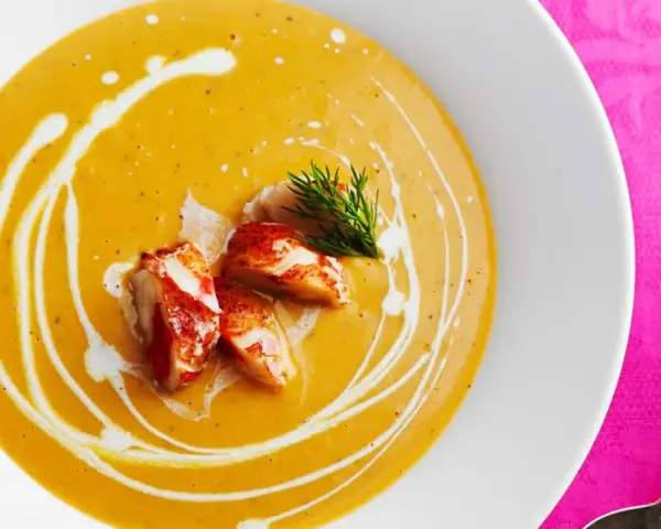 Un cremoso bisque de langosta, adornado con trozos de carne de langosta y un toque de eneldo, servido en un plato blanco sobre un mantel rosa vibrante.