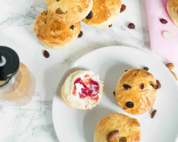 Goldbraune Scones mit Rosinen und Mandeln, ein Scones Teig mit Marmelade und Sahne, daneben ein Getränk.