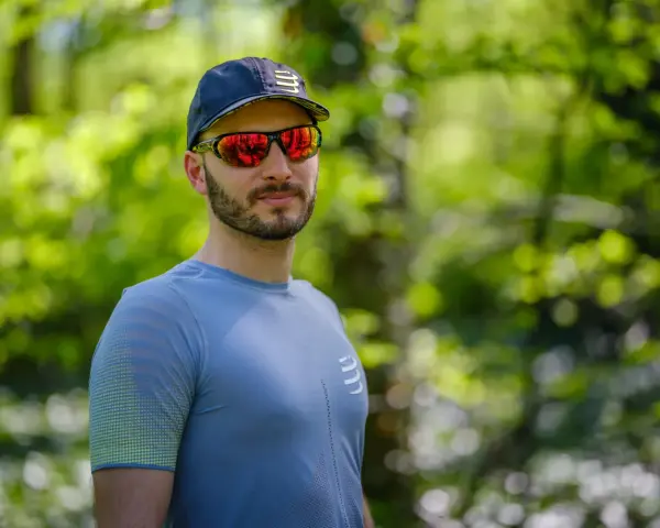 Mężczyzna w czapce i sportowych okularach przeciwsłonecznych, z odbiciem lasu w soczewkach.
