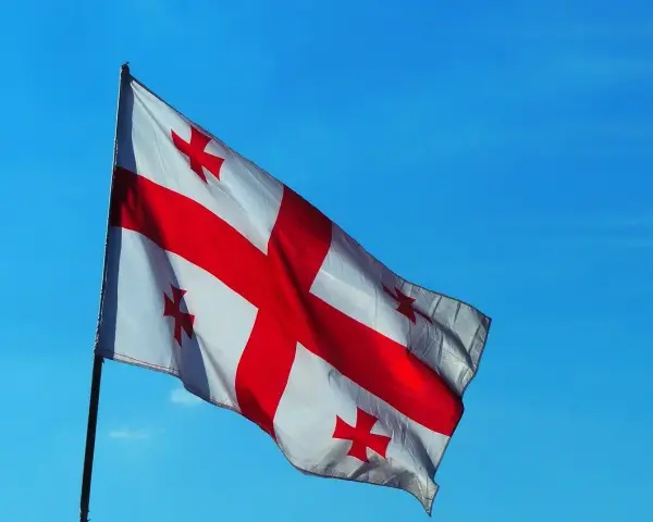 Flaga Gruzji powiewa na tle błękitnego nieba. Białe tło przecina czerwony krzyż, a w każdym z czterech pól znajduje się mniejszy czerwony krzyżyk.