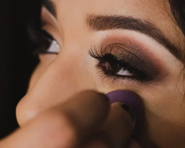 Un œil maquillé avec un smoky eyes facile, rehaussé de paillettes. Une éponge violette est utilisée pour estomper le fond de teint.