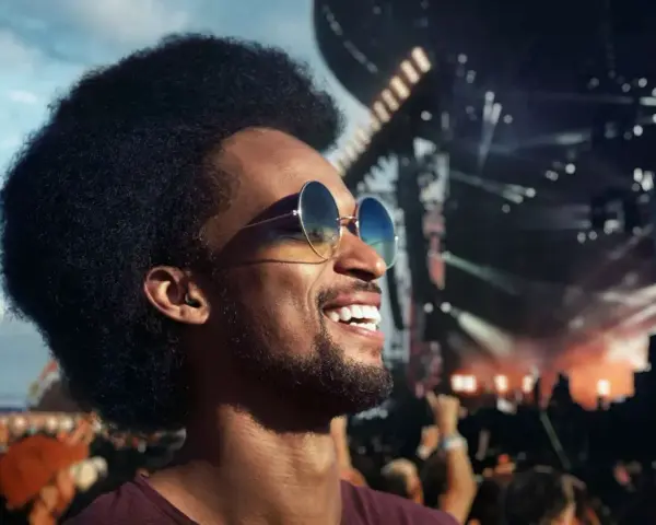 Un homme souriant avec des lunettes de soleil et des bouchons d'oreilles, profitant d'un concert en plein air. Le port des bouchons d'oreilles est obligatoire pour protéger son audition.