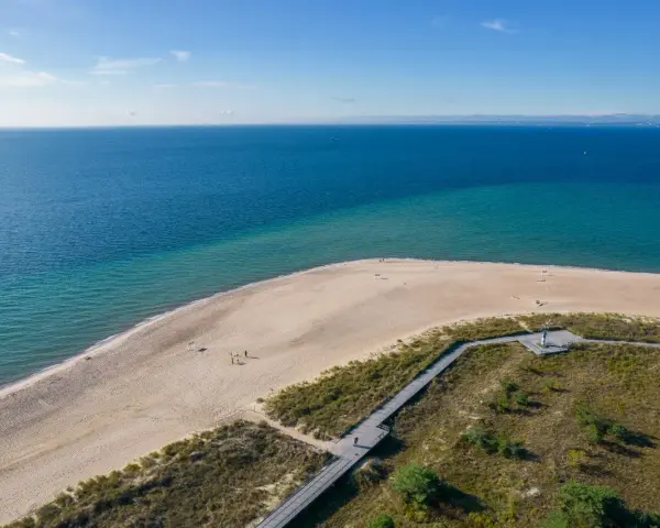 Piękne wydmy helskie nad turkusowym morzem. Drewniana kładka prowadzi przez zarośla na piaszczystą plażę, gdzie spacerują nieliczni turyści.
