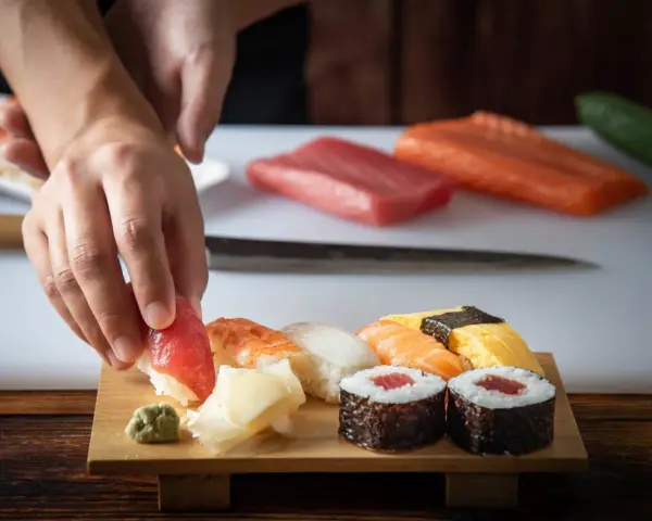 Ręce układają kawałek tuńczyka na ryżu, tworząc sushi. Na desce widać też krewetkę, kalmara, łososia i tamago. To inspiracja, jak zrobić sushi.