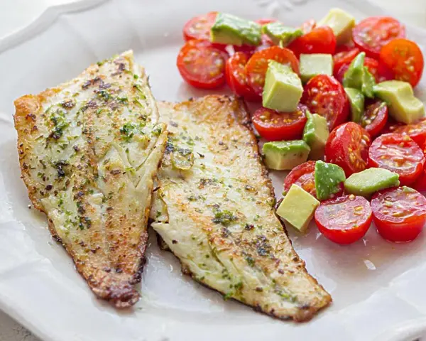 Dos filetes de lubina a la plancha con hierbas, acompañados de una ensalada fresca de tomates cherry y aguacate en cubos.