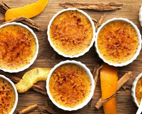 Flera krämiga crema catalana i vita skålar, toppade med karamelliserat socker. Apelsinskal och kanelstänger ger en härlig doft.