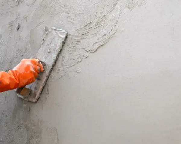 Main d'un ouvrier en gant orange appliquant de l'enduit sur un mur avec une truelle. Apprentissage de comment faire un enduit sur un mur.