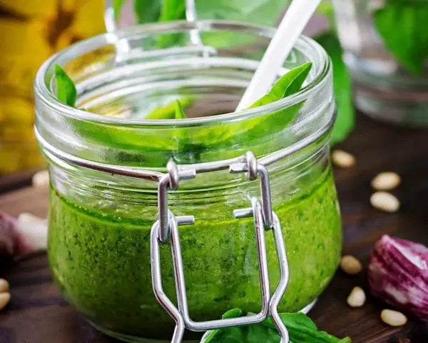 Un pot de pesto ail des ours, vert vif, avec une cuillère blanche. Des pignons de pin et de l'ail parsèment la table en bois.