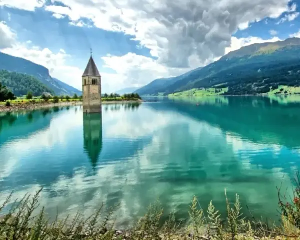 Il lago del campanile, con la sua torre sommersa, riflette montagne verdi e un cielo nuvoloso.