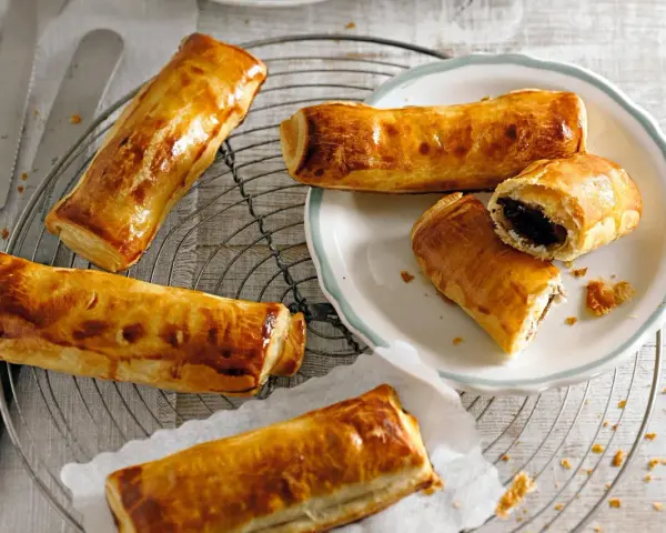 Goldbraune, knusprige Pain au Chocolat auf einem Gitter, einige auf einem Teller, einer aufgeschnitten.