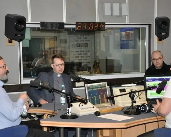 W studiu radiowym czterech mężczyzn, w tym ksiądz i osoba w jarmułce, rozmawia przy mikrofonach. W tle widać zegar i logo RDC.