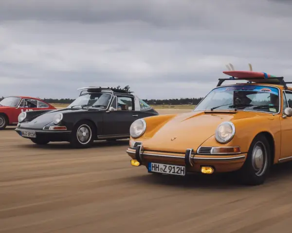 Trzy klasyczne Porsche jadące po plaży. Zdjecie w ruchu, z deskami surfingowymi na dachu jednego z nich.