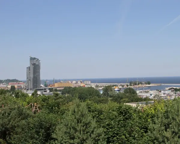 Widok na port w Gdyni z dominującym wieżowcem. W oddali morze, a na pierwszym planie zieleń drzew. Kamienna Góra w Gdyni oferuje takie panoramy.