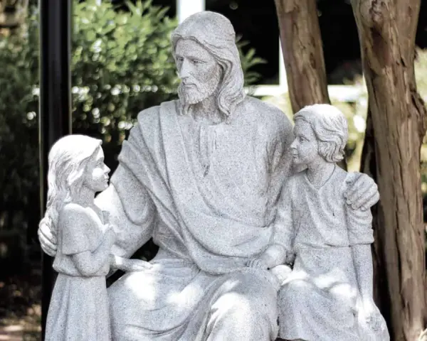 Steinfigur von Jesus, der zwei Kinder umarmt. Er wirkt wie ein liebevoller Gottvater, der seine Schützlinge beschützt.