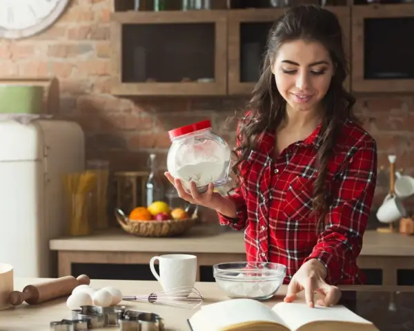 Junge Frau liest Rezept und hält Mehl bereit. Sie überlegt, ob sie Wasser statt Milch beim Backen verwenden soll.