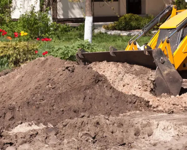 Koparka z łyżką zasypuje ziemię, przygotowując teren pod budowę. W tle kwitnące tulipany.