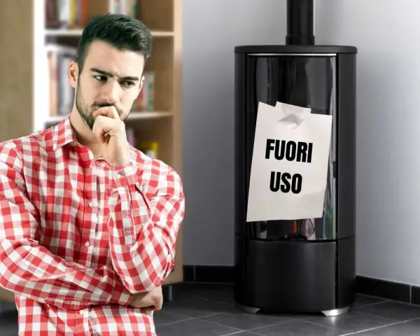Uomo pensieroso guarda stufa spenta con foglio "FUORI USO". Necessaria riparazione stufe per il freddo.