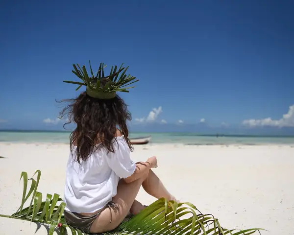 Kobieta w koronie z liści siedzi na plaży, marząc o tym, jak dolecieć na Zanzibar. Błękitne niebo i turkusowa woda zapraszają do podróży.