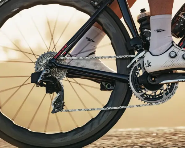 Close-up of a cyclist's shoe and drivetrain, showing the chain, gears, and crankset. The focus is on the power transfer, hinting at the cycling watts being generated.