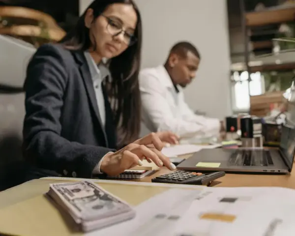 Kobieta w okularach liczy na kalkulatorze, obok stos banknotów i laptop.