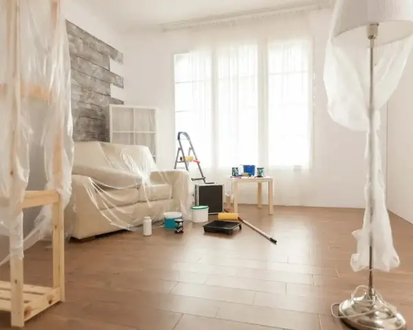 Przygotowania do malowania paneli podłogowych. Meble i lampa przykryte folią, na podłodze wałek i kuweta z farbą.