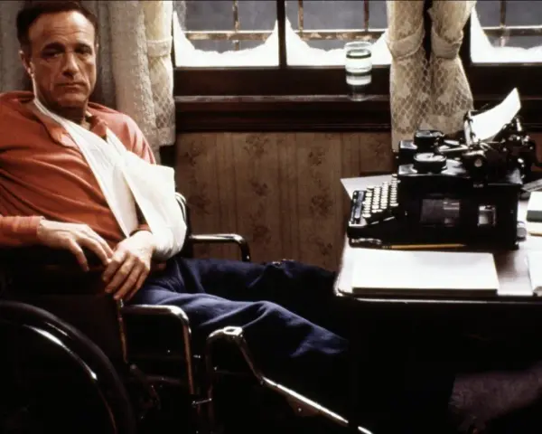 Un escritor en silla de ruedas, con un brazo en cabestrillo, mira fijamente una máquina de escribir. Escenas como esta son comunes en películas sobre escritores.