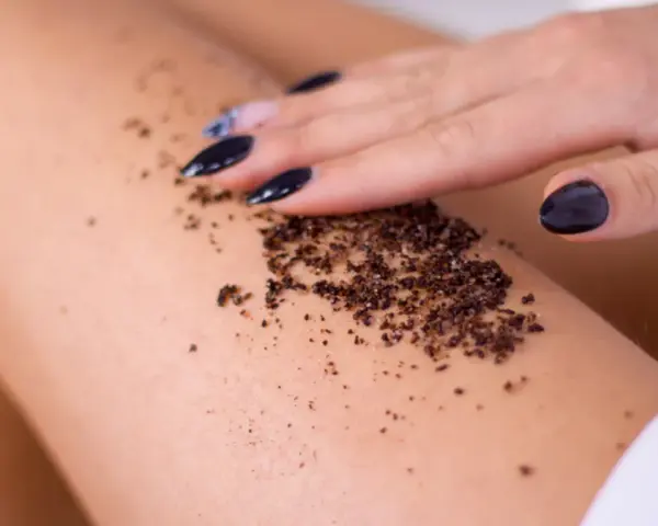 Dłoń z czarnym lakierem rozprowadza na skórze odżywczy peeling z kawy, przygotowując ją na letnie słońce.