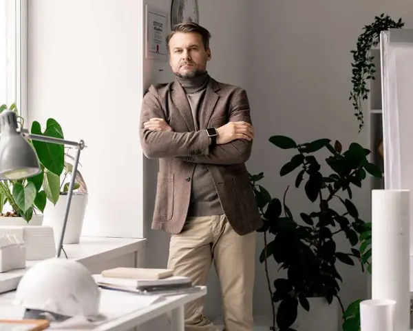 Mężczyzna w marynarce stoi przy oknie, obok roślin. Na biurku leży biały kask, a w tle widać projekt. Prawdopodobnie zajmuje się **operat wodnoprawny**.