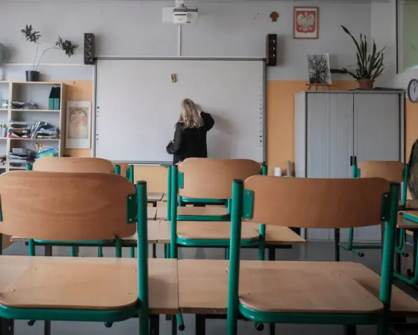 Nauczycielka przy tablicy, zastanawia się, ile zarabia przedszkolanka. Puste ławki w sali lekcyjnej.