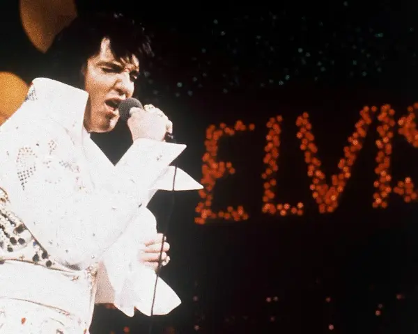 Elvis Presley singt in seinem berühmten weißen Anzug in Las Vegas, während sein Name in roten Lichtern erstrahlt.