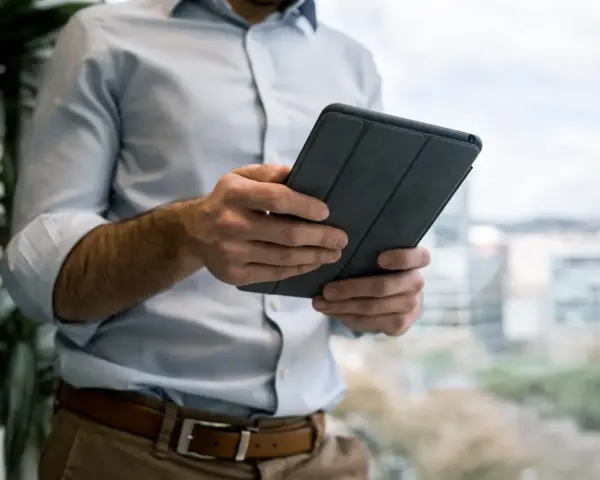 Mężczyzna w jasnej koszuli i brązowych spodniach trzyma tablet w szarej okładce. e cmr.