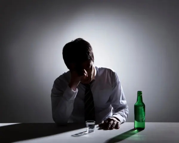 Mężczyzna w białej koszuli i krawacie siedzi przy stole, trzymając się za głowę. Obok stoi szklanka i zielona butelka. To obraz przedawkowania alkoholu.