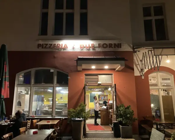 Abendstimmung vor der Pizzeria "Due Forni". Ein Mann steht im Eingangsbereich, während Gäste an Tischen im Freien sitzen.