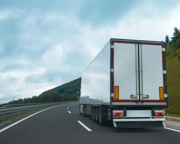Ciężarówka na autostradzie, z tyłu widoczne białe drzwi naczepy. W tle góry i chmury. Tablice wyróżniające przepisy są częścią oznakowania pojazdu.