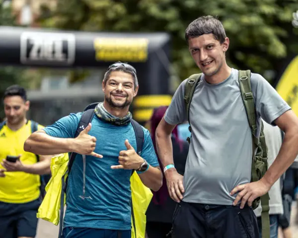 Zwei Männer mit Rucksäcken, einer mit Daumen hoch, der andere mit Handzeichen. Sie sind bereit für ihr Abenteuer, bei dem die richtige **Merinowolle Pflege** wichtig ist.