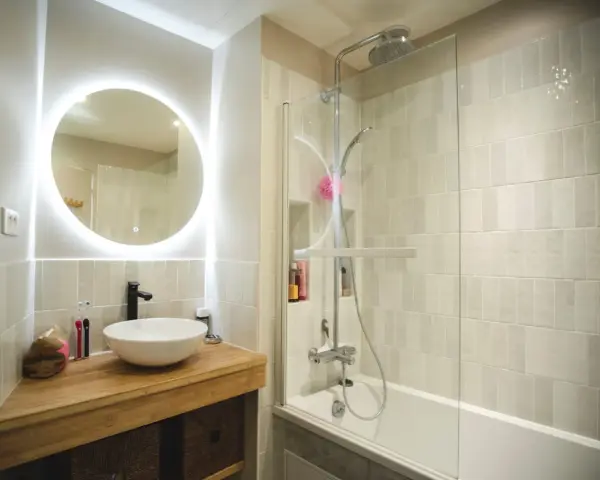 Une **salle de bain blanche** moderne avec un lavabo rond sur un meuble en bois, un miroir lumineux et une douche avec carrelage clair.