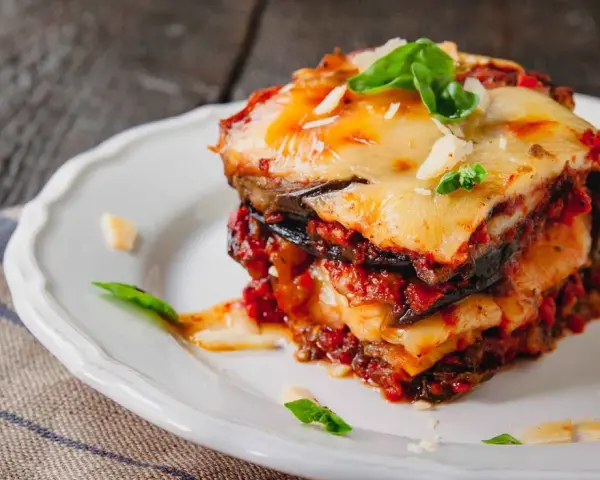 Una fetta di parmigiana di melanzane al forno, stratificata con melanzane, sugo e formaggio filante, guarnita con basilico fresco.