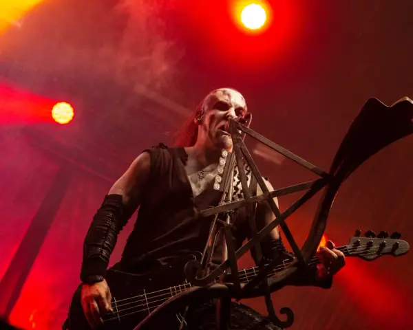 Un musicien au visage peint, jouant de la basse sur scène, baigné de lumières rouges. Sa posture exprime une vie d'agonie.