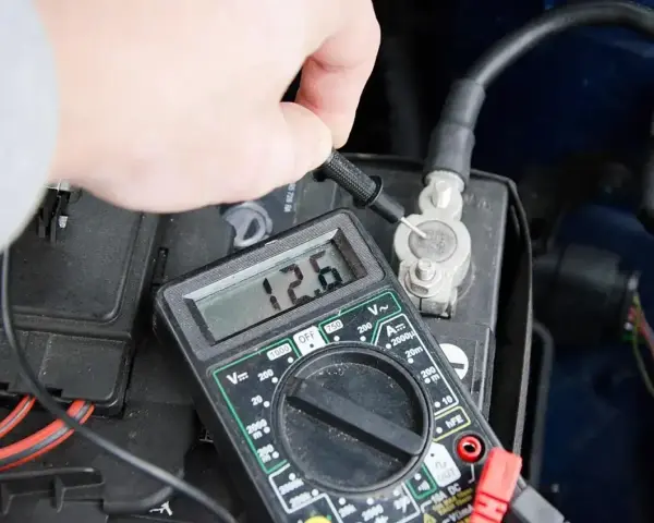 Dłoń mierzy napięcie akumulatora multimetrem. Wynik 12.6V. Widoczna jest jedna z klem.