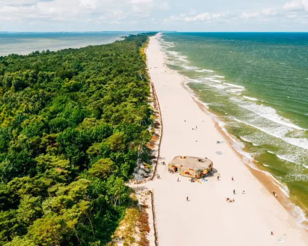 Majówka nad morzem: zielony las, piaszczysta plaża z kilkoma osobami i bunkrem w kamuflażu, szmaragdowe fale Bałtyku.