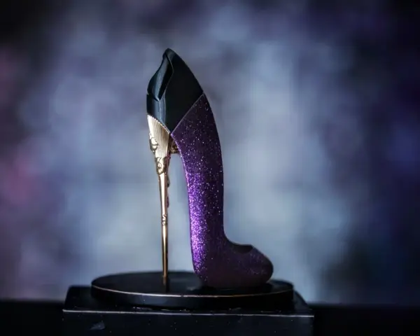 Elegancki perfumy bucik w kolorze fioletowym, ozdobiony złotymi detalami i brokatem, stoi na czarnym postumencie.