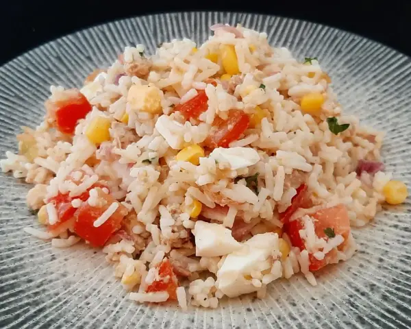 Une généreuse portion de salade de riz thon, garnie de dés de tomates, de maïs et d'œufs durs, sur une assiette texturée.