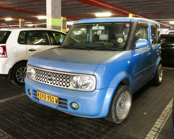 Niebieski Nissan Cube z charakterystycznym grillem i tablicą rejestracyjną N193-701 W, zaparkowany obok białego Volkswagena.