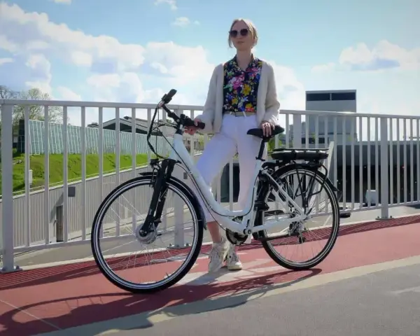 Kobieta w białych spodniach i kolorowej koszuli stoi obok białego roweru elektrycznego. Czy na rower elektryczny trzeba mieć prawo jazdy?