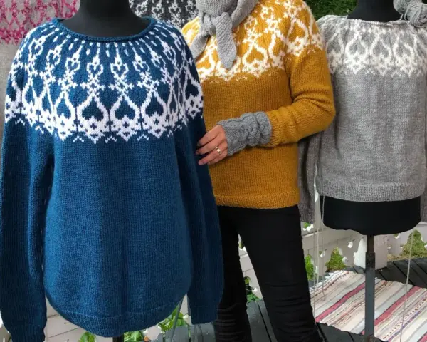Tre stickade tröjor med mönster, en blå, en gul och en grå. En av dem visas på en docka, redo att sticka tröja nerifrån och upp på rundsticka.